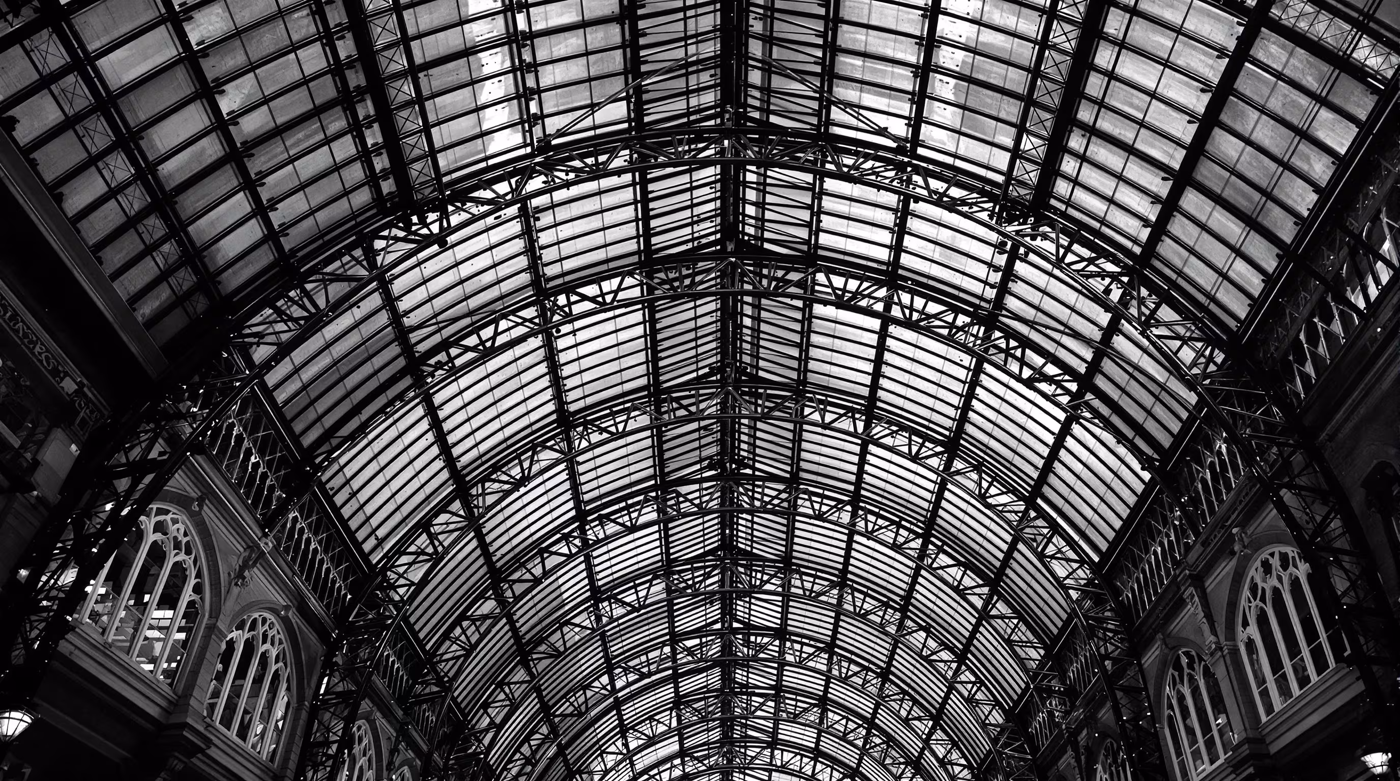 A Victorian iron and glass arcade ceiling at night, raking light across the tracery.